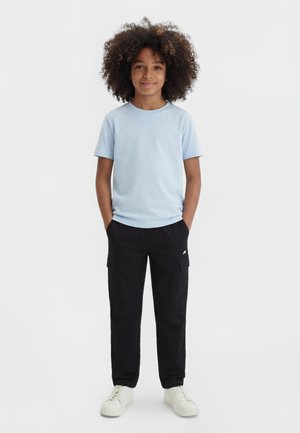 Jeune personne aux cheveux bouclés souriante, portant un t-shirt bleu clair, un pantalon cargo noir et des baskets blanches sur un fond uni.
