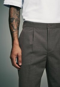 Gray pinstriped trousers with pleats, featuring a button closure and a sleek, smooth fabric texture. Hand with visible tattoo resting nearby.