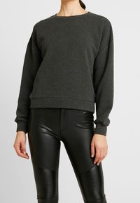 Sudadera de manga larga de canalé gris oscuro con cuello redondo. Combinada con pantalones ajustados negros que tienen acentos texturizados y un acabado brillante.