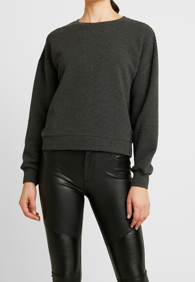 Sudadera de manga larga de canalé gris oscuro con cuello redondo. Combinada con pantalones ajustados negros que tienen acentos texturizados y un acabado brillante.