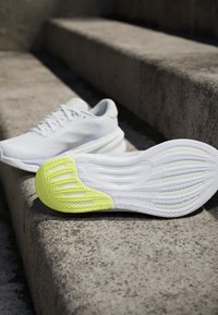 Witte sportschoenen die op betonnen trappen liggen, waarbij één schoen een heldere gele, gestructureerde zool naar boven toont.