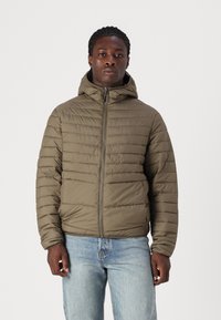 Olivgrüne Daunenjacke mit Kapuze, horizontal gestepptem Design, Frontreißverschluss und elastischen Bündchen. Darüber werden hellblaue Jeans getragen.