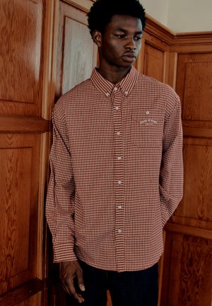 Jeune homme portant une chemise à carreaux rouge et blanche, debout devant des murs en bois, regardant vers le bas et sur le côté.