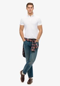 Vit kortärmad polo, blå jeans, bruna skor, rutig skjorta knuten runt midjan, klocka på handleden, stående pose med armarna i kors.