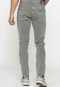 Jean en denim gris avec une coupe ajustée, comportant deux poches arrière et une étiquette sur la taille. Porté avec des chaussures de sport gris clair.