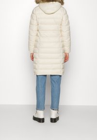Padded white coat with a hood and faux fur trim, featuring a straight cut, long sleeves, and a smooth texture. Paired with light blue jeans and white ankle boots.