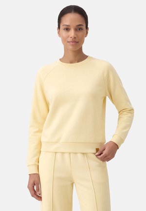 Vrouw die een lichtgele lange mouwen trui en bijpassende broek draagt, staand tegen een effen lichte achtergrond.