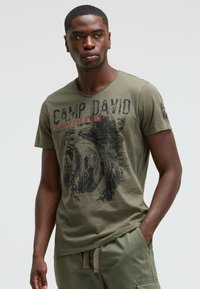 Olive green cotton T-shirt with a v-neck, featuring black and red graphic print that includes text and designs. Short sleeves, relaxed fit.