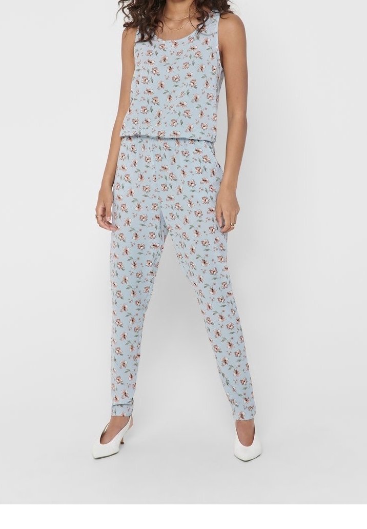Femme portant un haut à fleurs sans manches bleu clair et un pantalon assorti, associé à des talons blancs à bouts pointus, debout devant un fond uni.