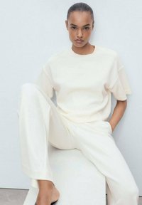 T-shirt rilassato di colore crema e pantaloni coordinati in un tessuto morbido. L'outfit presenta un design semplice con spalle scese e accenti minimi.