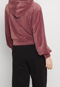 Felpa corta con cappuccio in un morbido e soffice tessuto mauve. Presenta maniche ampie con polsini arricciati e un design semplice. Abbinata a pantaloni scuri.