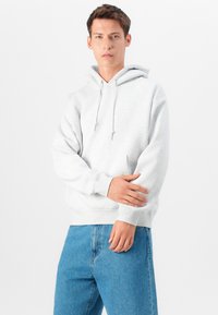 Camisola capuz cinza com um capuz ajustável, bolso canguru, punhos e bainha canelados. Usada com jeans denim azuis. Tecido suave e texturizado.