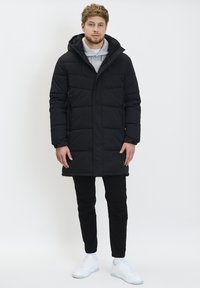 Sort pufferjakke med hætte, med en quiltet design, sidelommer og en afslappet pasform, parret med sorte bukser og hvide sneakers.