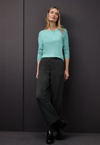 Turkos långärmad topp, svarta texturerade vida byxor och svarta ankelstövlar. Modellen står framför en mörkgrå bakgrund.