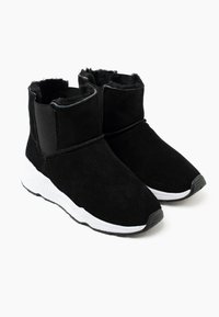 Botines de ante negro con suela de goma blanca, paneles laterales elásticos y forro interior suave. Puntera redondeada y un diseño minimalista.