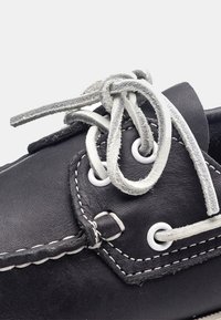 Zapato de barco negro de cuero con costuras y ojales blancos. Presenta cordones grises con un nudo. Punta redondeada y superficie texturizada.