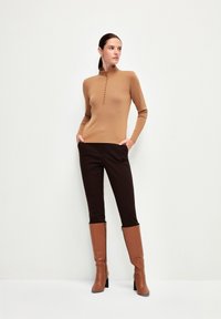 Hellbraunes, figurbetontes Sweater mit hohem Kragen und Knopfdetails, kombiniert mit dunkelbraunen Hosen und hohen, braunen Stiefeln mit Absatz.