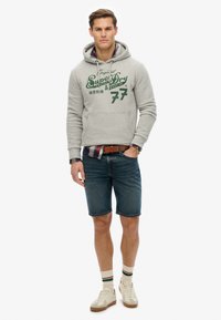Grå hoodie med grøn logo, parret med blå denimshorts, hvide sneakers og stribede sokker. Modellen står foran en ensfarvet baggrund.
