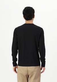 Black long-sleeve shirt with subtle embossed pattern, crew neckline, and straight cut. Fabric appears soft, worn with beige pants.