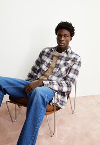 Gestrikt shirt in blauw en wit met bruine tinten, gecombineerd met een gestructureerde beige trui en denim jeans, zittend op een metalen stoel.