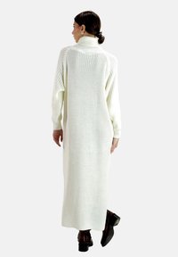 Lang, hvid strikket sweaterkjole med høj krave, ribbet tekstur, nedsatte skuldre og en afslappet pasform. Bæres med brune hælede støvler.
