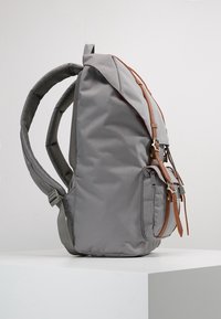 Le sac à dos en tissu gris présente un design simple, des bretelles ajustables et une poche frontale mise en valeur par un accent en cuir brun.