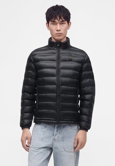 Homme portant une veste matelassée noire avec fermeture éclair, debout devant un fond clair uni, associée à un jean bleu clair.