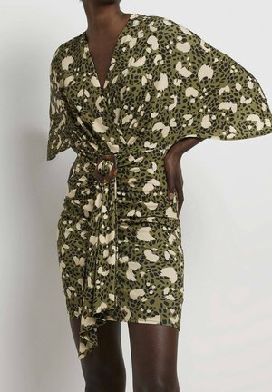 Kvinde iført kort olivengrøn kjole med sorte og hvide abstrakte pletter, vide ærmer og et stofbælte med en brun spænde.