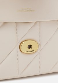 Jasno beżowa pikowana skórzana torba z złotą owalną klamrą obrotową z wygrawerowanym napisem "COACH" oraz wytłoczonym napisem "THE TABBY BAG GENUINE NAPPA LEATHER" powyżej.