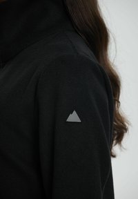 Sort fleecejakke med høj krave og et trekantet sølvbjergmærke på venstre skulder. Blød tekstur, tætsiddende design.