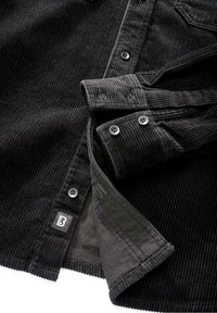 Chemise en velours côtelé noir avec fermeture à boutons sur le devant, manches longues, et étiquette sur l'ourlet. Texture côtelée marquée et boutons contrastants.