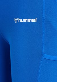 Blåt atletisk stof med hvidt "hummel" logo og bi-ikon, med detaljerede syninger og en netlomme på siden.