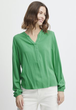 Blouse verte à manches longues en tissu texturé, avec un col en V et des poignets élastiques. Elle a une coupe décontractée et un dos légèrement plus long.