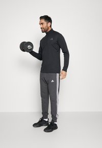 Črna zip-up športna majica z diskretnim logotipom, v kombinaciji s sivo trenirka s belimi stranskimi črtami in črnimi športnimi čevlji, medtem ko drži uteži.