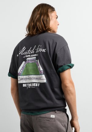 Mann mit schulterlangem Haar, der ein schwarzes T-Shirt mit einer farbenfrohen Stadiongrafik und sportlichem Text auf der Rückseite trägt.