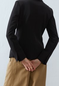 Mustat räätälöidyt bleiserit pitkillä hihoilla, sileä pinta, vartalonmyötäinen muotoilu ja levenevät hihansuut, yhdistettynä beigeihin korkeavyötäröisiin housuihin.