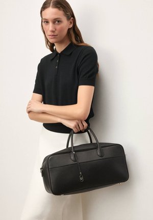 Geantă din piele neagră cu suprafață texturată, mânere rotunjite duble, accesorii argintii și închidere cu fermoar; purtată de un model îmbrăcat într-un tricou polo închis la culoare.