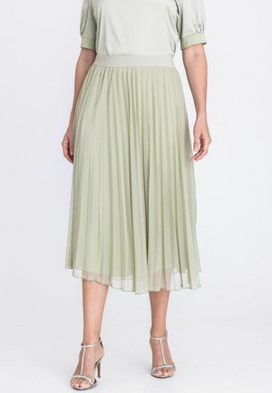 Femme portant une jupe midi plissée vert clair, un haut à manches courtes et des sandales à talons hauts argentées, debout devant un fond blanc.