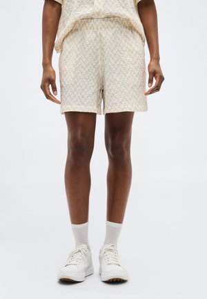 Inimene, kes kannab tekstuurse pindalaga beežikaid lühikesi pükse, ühesugust pealset, valgeid pahkluu sokke ja valgeid tenniseid, seisab ühevärvilise tausta ees.