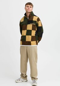 Fleece-pullover med hög krage, med ett lapptäckemönster i grönt, brunt och gult. Dragkedjeficka på bröstet, mjuk textur.