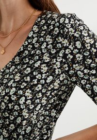 Czarna sukienka w kwiaty z małymi białymi i zielonymi kwiatami, krótkie rękawy oraz dekolt w kształcie litery V. Miękka tkanina o teksturowanym wykończeniu. Złoty naszyjnik jako dodatek.