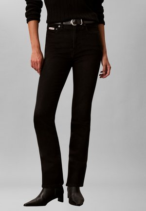 Person iført sorte jeans med lige ben, sort bælte, sorte spidse støvler og en sort ribstrikket sweater med ærmerne rullet op.