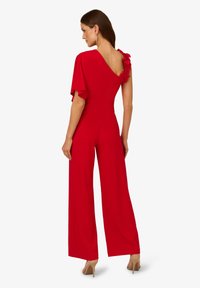 Červený overal s přiléhavým živůtkem, jedním volným rukávem a květinovými akcenty na rameni. Široké nohavice, hladký materiál a bezháčkový detail na zádech.