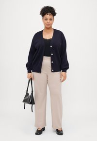 Cardigan azul-marinho com fecho de botão, top preto, calças largas bege, bolsa preta e sapatos fechados pretos. Cabelo encaracolado.