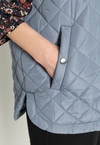 Prošívaná šedá vesta s boční kapsou a stříbrným patentním knoflíkem. Materiál má hladkou strukturu s diamantovými vzory.