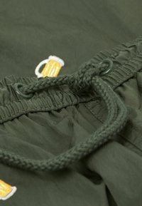 Zelené látkové kraťasy s elastickým pasem a šňůrkou. Má všité vyšívané logo piva. Hladký povrch a neformální design.