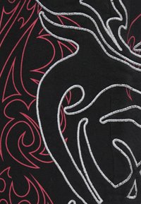 Μαύρο ύφασμα με περίτεχνη ασημένια και κόκκινη κεντήματα που παρουσιάζουν αφηρημένα στροβιλισμένα σχέδια και αιχμηρά μοτίβα. Ορατή είναι η υφή της επιφάνειας.