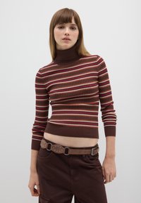 Braun gestreifter Rollkragenpullover mit gerippter Textur, versehen mit roten, orangen und cremefarbenen Linien, kombiniert mit braunen Hosen und einem Nieten-Gürtel.