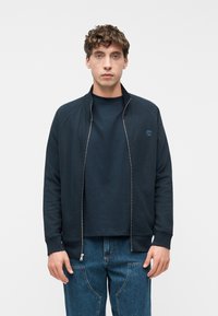 Νέος άνδρας με κατσαρά μαλλιά φοράει σκούρο μπλε μπουφάν με φερμουάρ πάνω από ένα ταιριαστό σκούρο μπλουζάκι και μπλε τζιν, όρθιος μπροστά σε μια απλή φόντο.