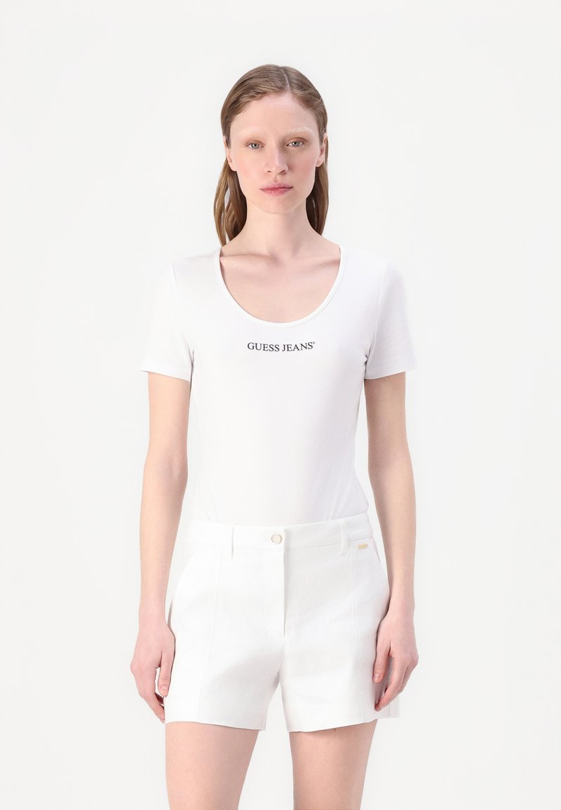 Camisa blanca de manga corta con escote redondo que presenta el logo "GUESS JEANS" en negro. Combinada con pantalones cortos blancos de corte ajustado con bolsillos frontales.